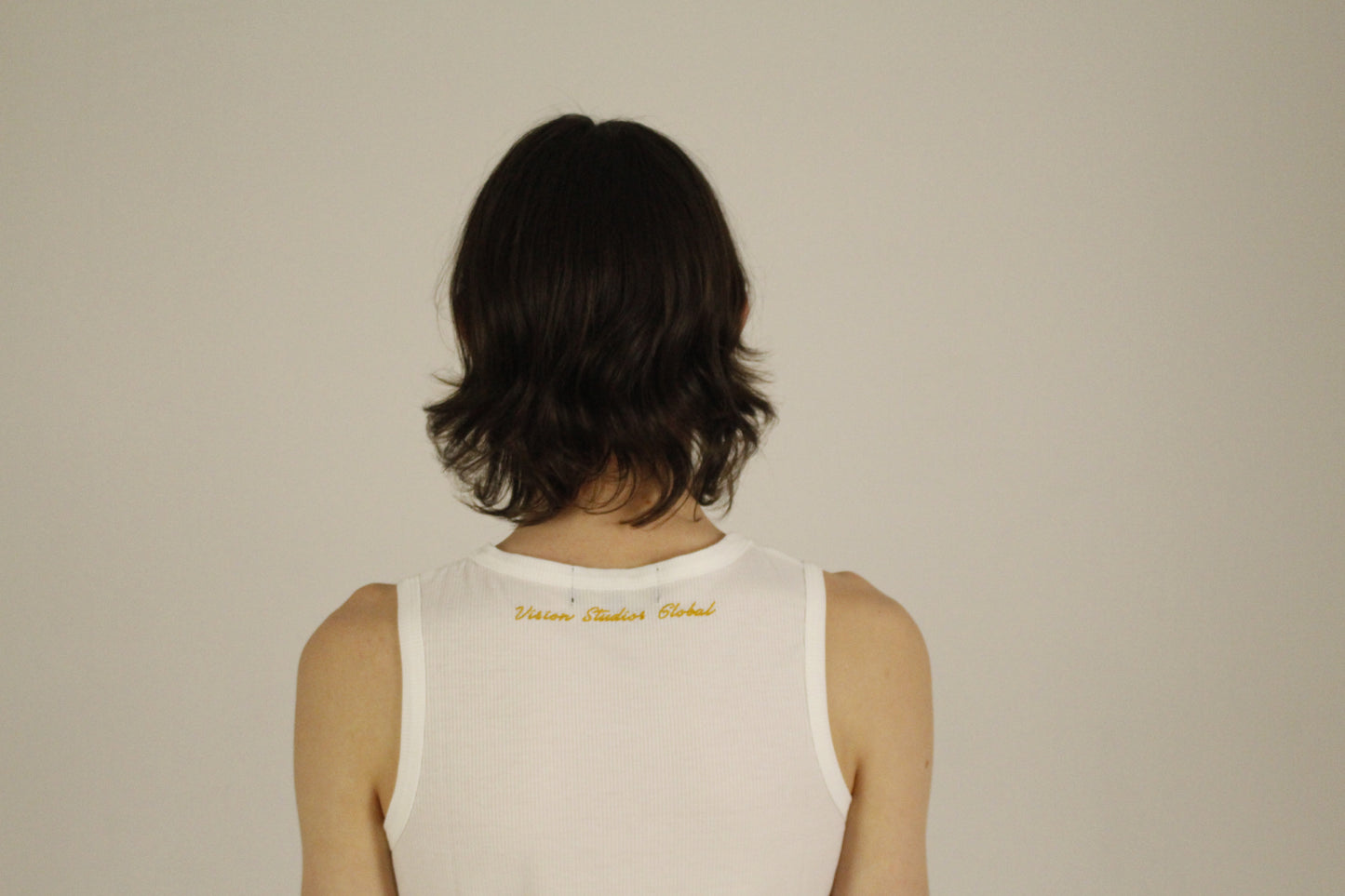 Back view of the VSG white ribbed tank top worn by a model, featuring a clean neckline and minimalist design. The neckline shows the golden embroidered Vision Studios Global logo.