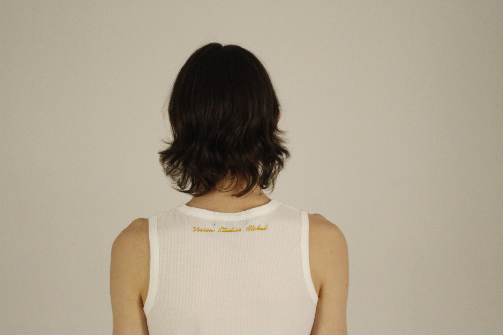 Back view of the VSG white ribbed tank top worn by a model, featuring a clean neckline and minimalist design. The neckline shows the golden embroidered Vision Studios Global logo.