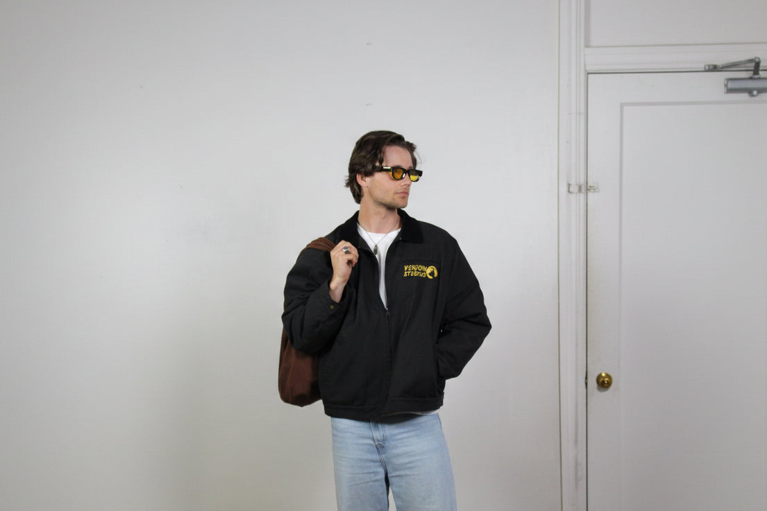 Man wearing a black jacket with a logo, holding a brown bag against a white background