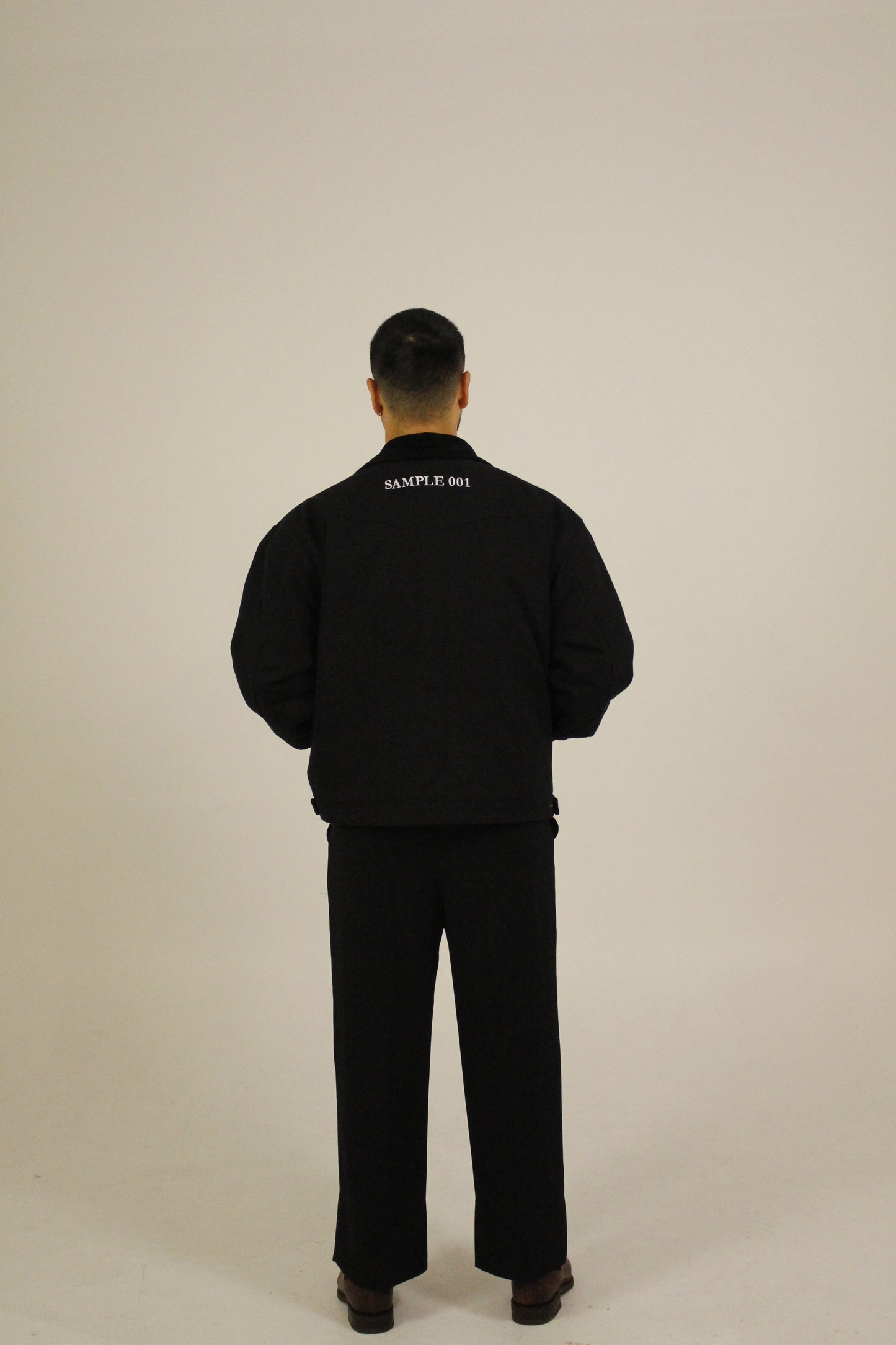 Back view of model wearing a black zip-up utility jacket featuring Vision Studios logo embroidery, styled with black pants in a studio setting