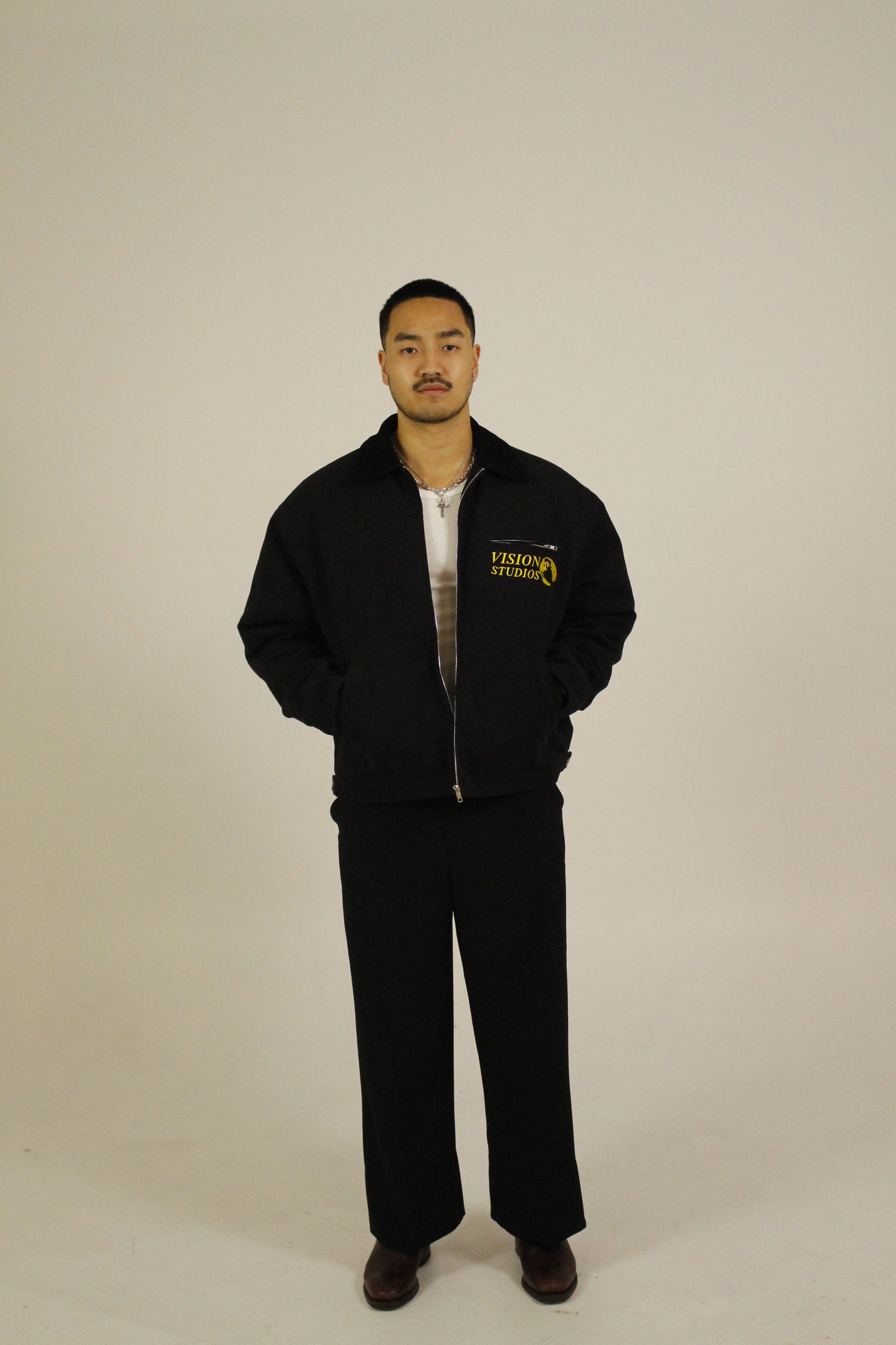 Front view of model wearing a black zip-up utility jacket unzipped featuring Vision Studios logo embroidery, styled with black pants in a studio setting