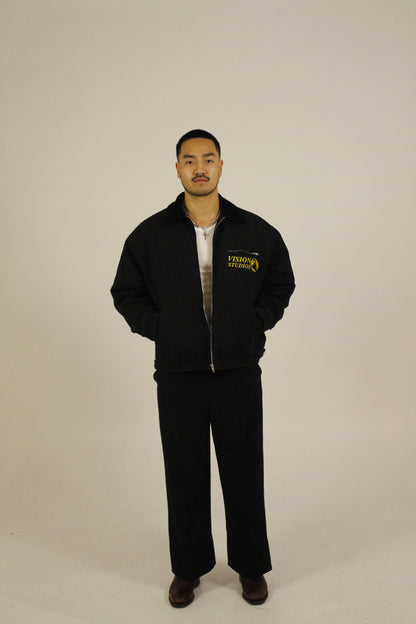Front view of model wearing a black zip-up utility jacket unzipped featuring Vision Studios logo embroidery, styled with black pants in a studio setting