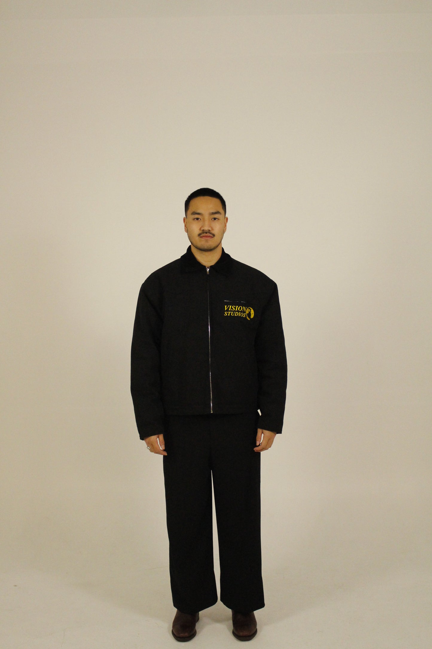 Front view of model wearing a black zip-up utility jacket featuring Vision Studios logo embroidery, styled with black pants in a studio setting
