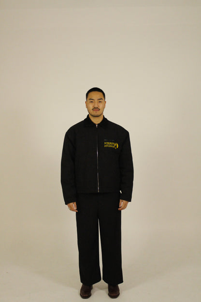 Front view of model wearing a black zip-up utility jacket featuring Vision Studios logo embroidery, styled with black pants in a studio setting