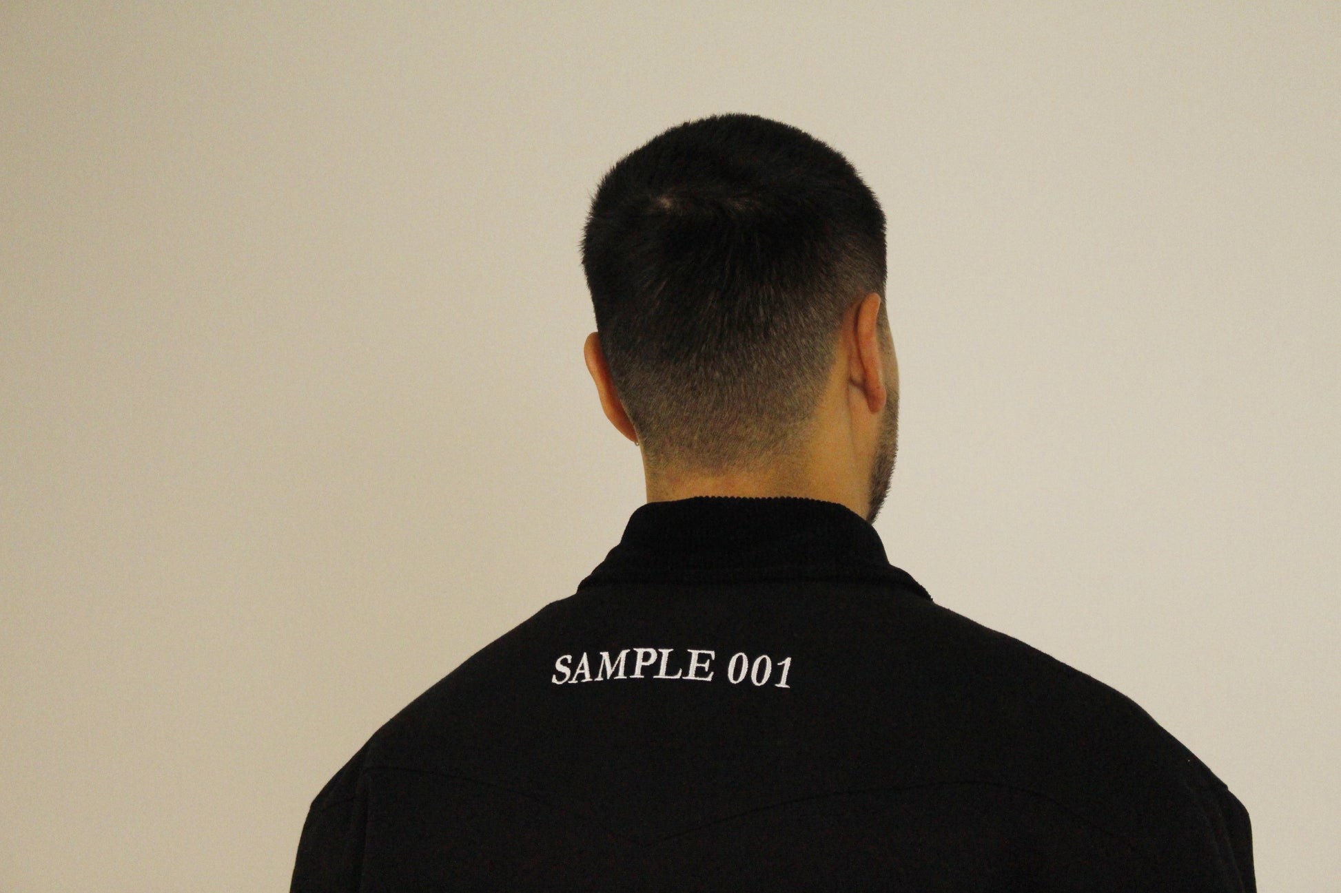 Close-up back view of black utility jacket showing SAMPLE 001 embroidered text on the collar neckline