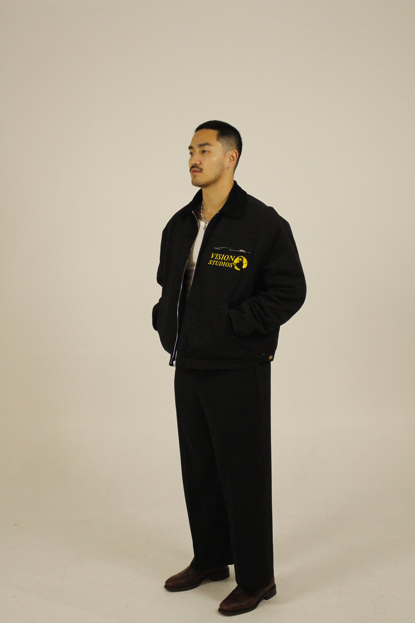 Side view of model wearing a black zip-up utility jacket unzipped featuring Vision Studios logo embroidery, styled with black pants in a studio setting