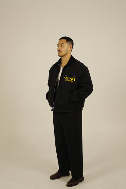 Side view of model wearing a black zip-up utility jacket unzipped featuring Vision Studios logo embroidery, styled with black pants in a studio setting