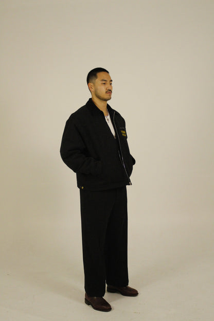 side view of model wearing a black zip-up utility jacket unzipped featuring Vision Studios logo embroidery, styled with black pants in a studio setting