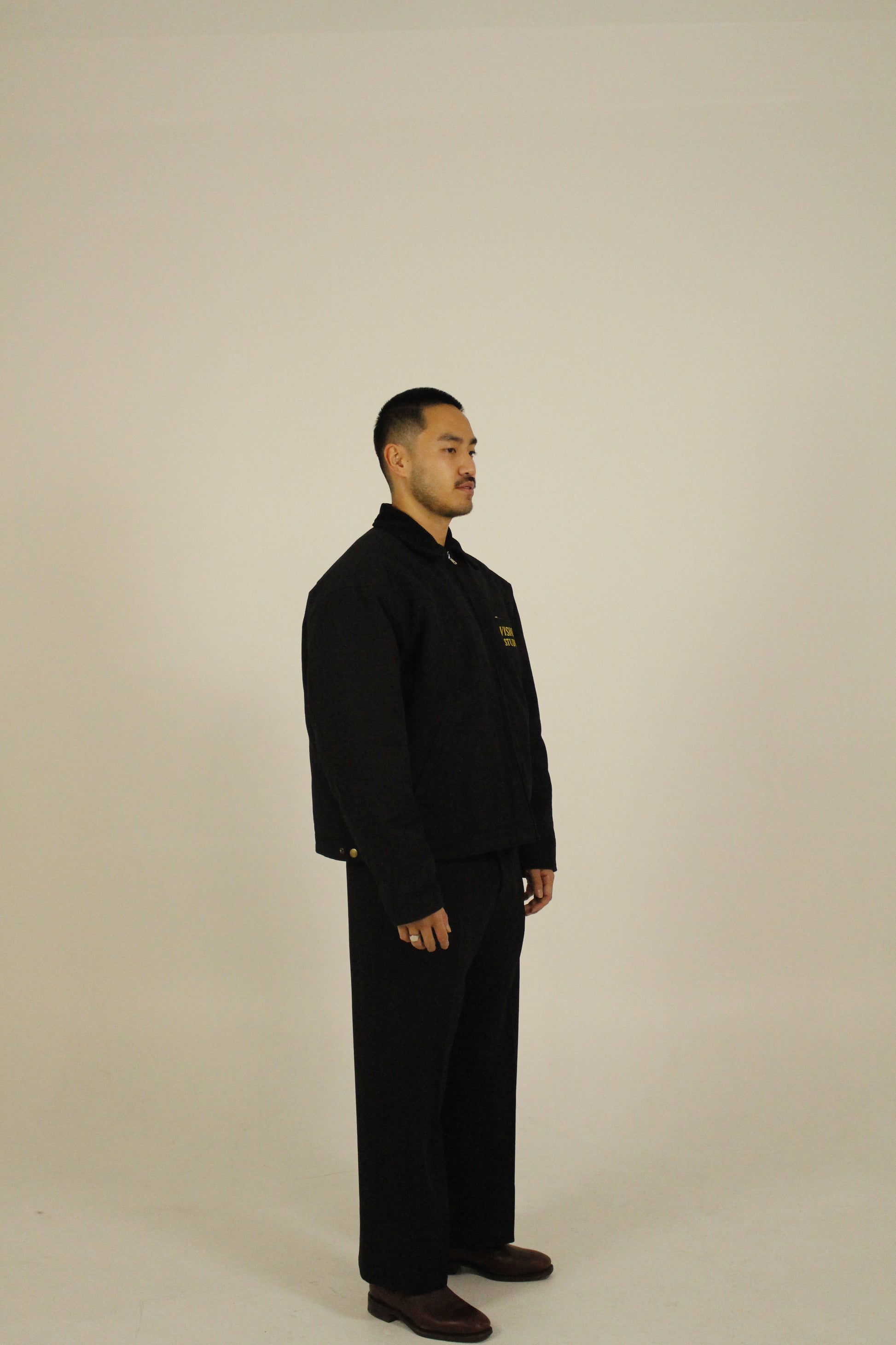 Side view of model wearing a black zip-up utility jacket featuring Vision Studios logo embroidery, styled with black pants in a studio setting