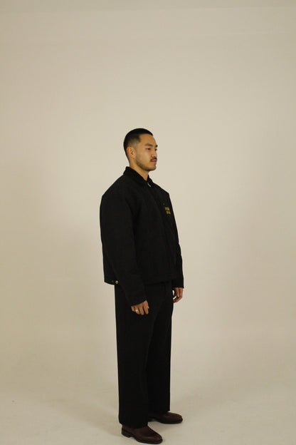 Side view of model wearing a black zip-up utility jacket featuring Vision Studios logo embroidery, styled with black pants in a studio setting