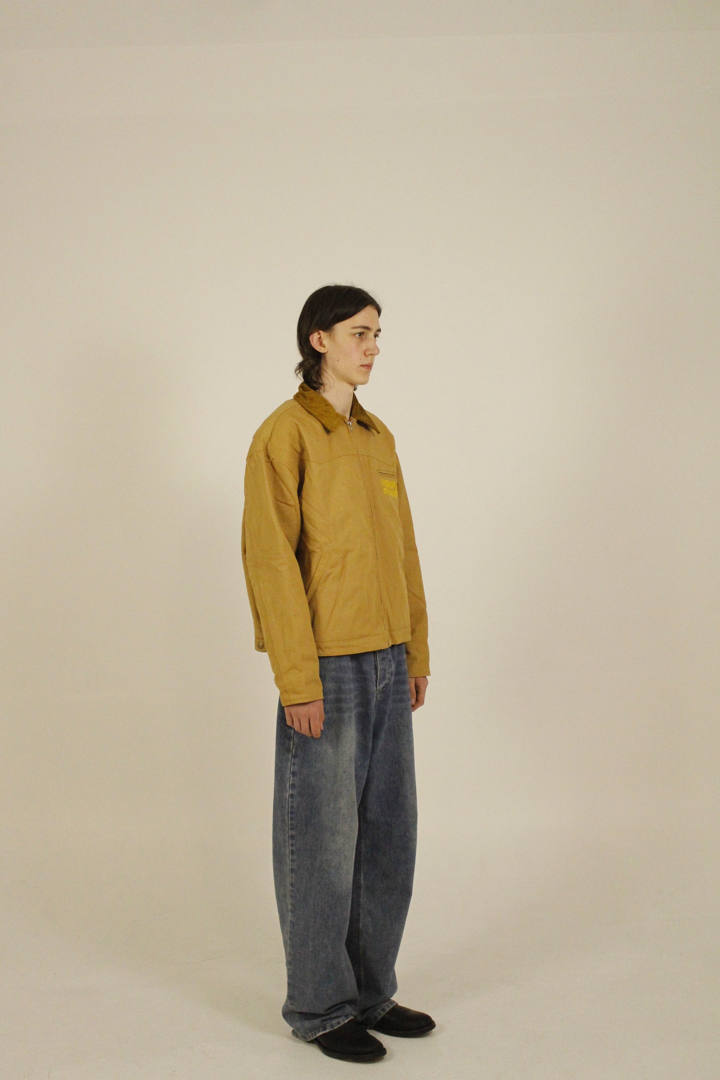 Side view of model wearing SAMPLE_001 canvas utility jacket zipped with embroidered Vision Studios logo, showing cropped workwear fit and structured silhouette