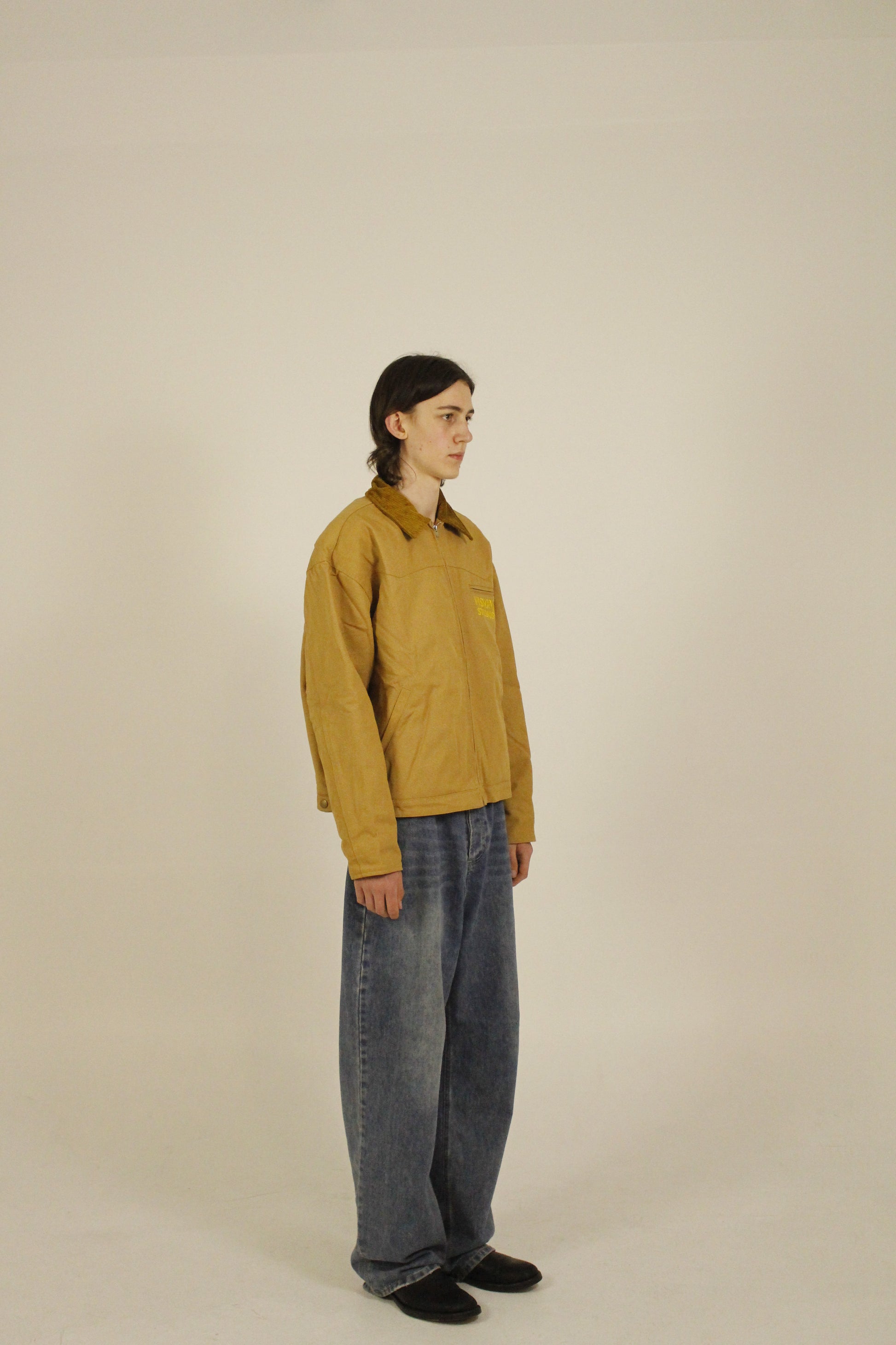 Side view of model wearing SAMPLE_001 canvas utility jacket zipped with embroidered Vision Studios logo, showing cropped workwear fit and structured silhouette