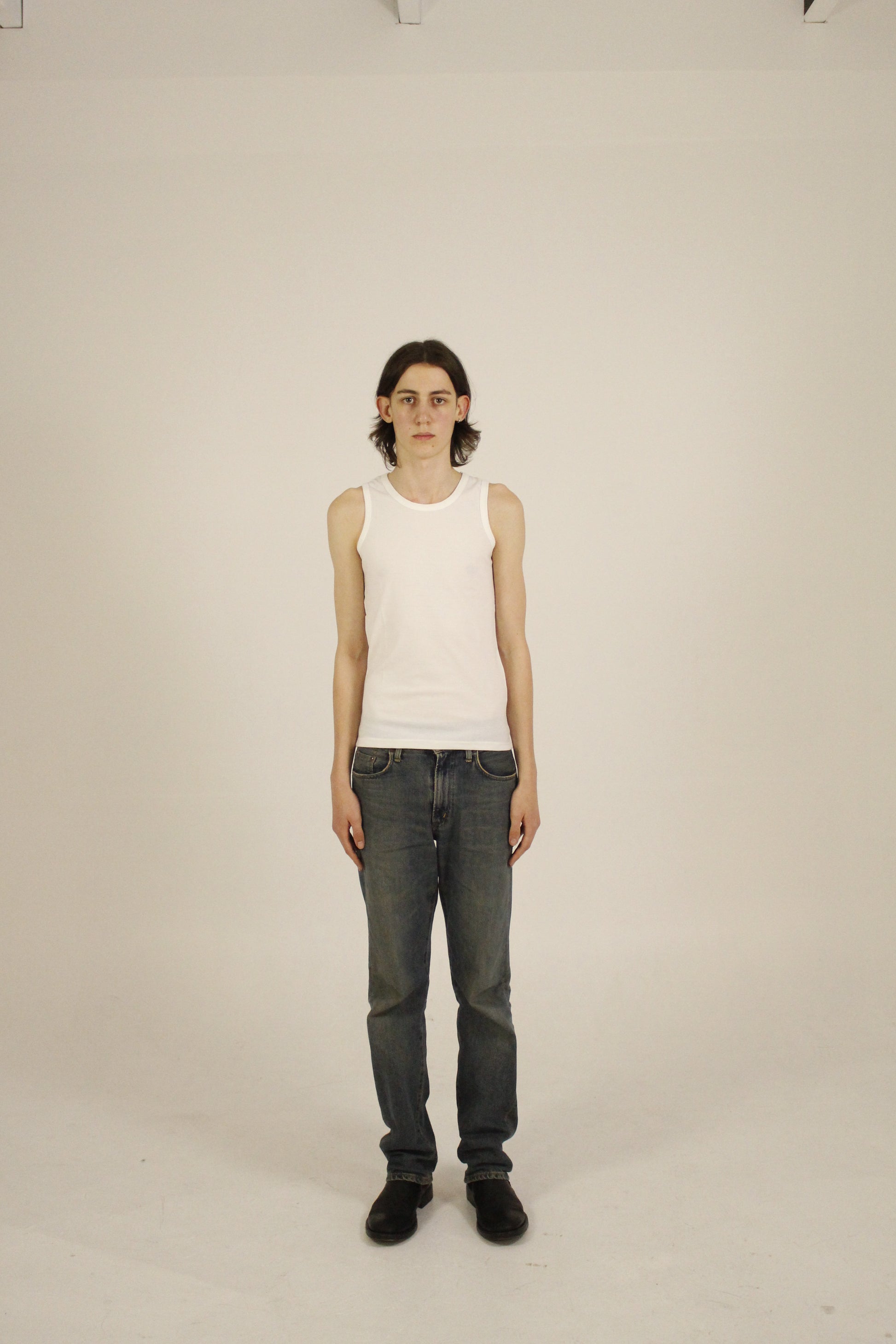 A person standing against a white background, wearing a ribbed tank top and dark pants, with a white tank top being displayed.