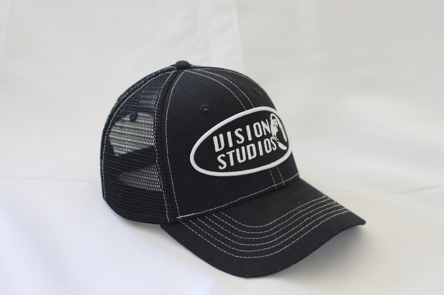 Black trucker hat with white stitching and vision studios logo on the front panel