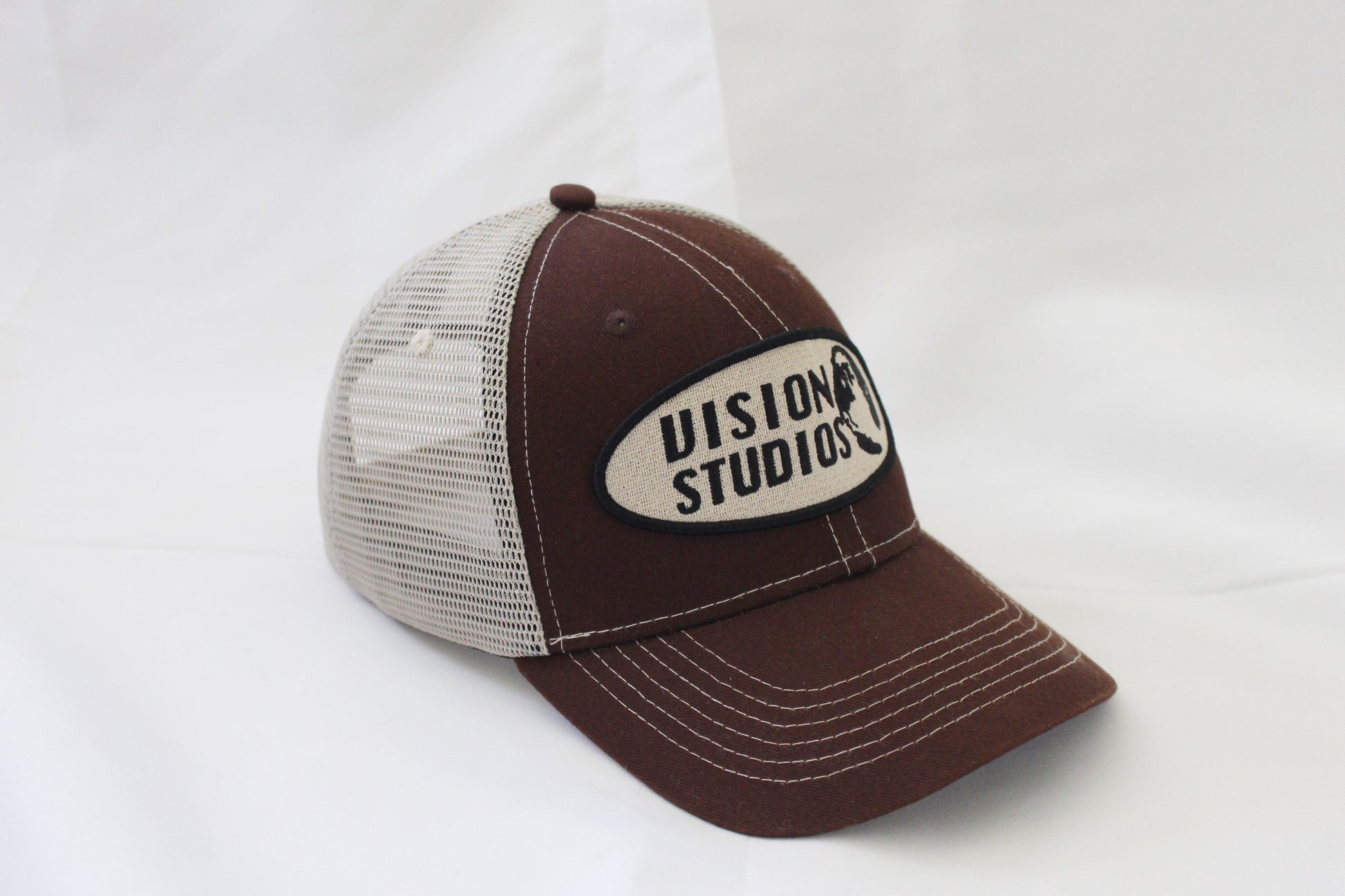 Brown trucker hat with white stitching and vision studios logo on the front panel