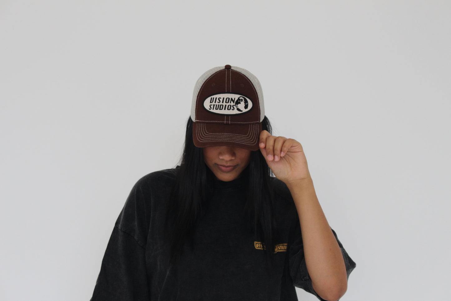 A female model wearing a posing with the brown vision studios trucker hat and acid black vision studios oversized tee