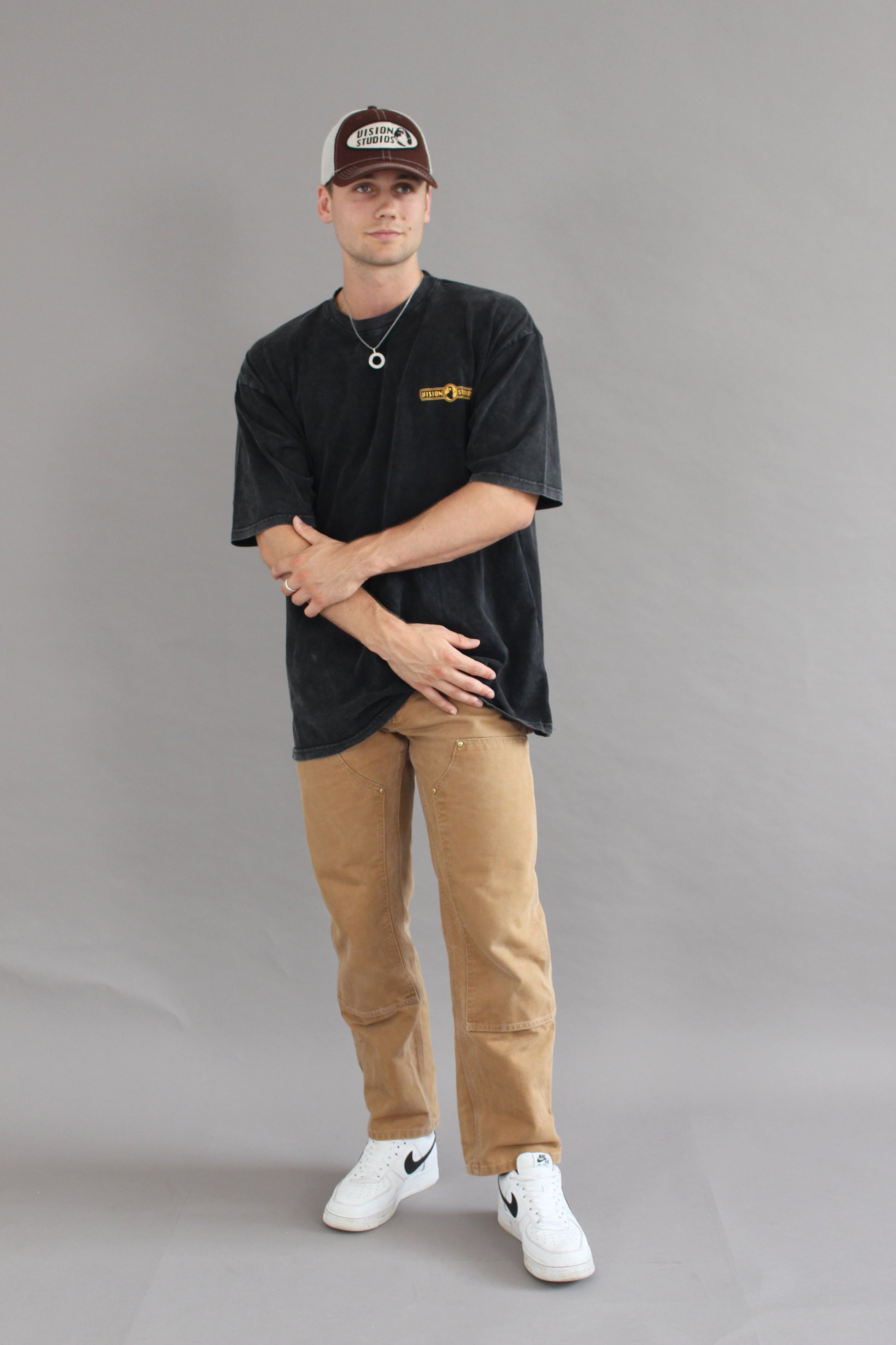 A male model wearing the brown vision studios trucker hat  and vision studios acid wash oversized tee